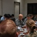 A formal military conference or briefing is taking place around a large wooden conference table. Personnel in multiple camouflage uniform patterns, including what appears to be USMC MARPAT and an Asian military digital camouflage pattern, are seated together alongside civilian attendees. The meeting appears to be a multinational military coordination session with documents, name placards, and beverages on the table.