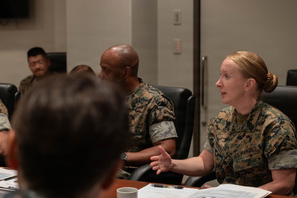 A group of U.S. Marines are seated around a conference table during what appears to be a briefing or meeting. A female Marine in MARPAT camouflage uniform is actively speaking and gesturing with her hand, appearing to be the primary presenter or discussant. Several other Marines in similar uniforms are seated nearby, with documents and a coffee cup visible on the table.