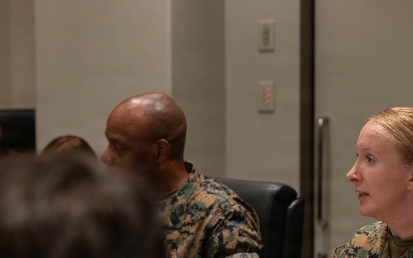 A group of U.S. Marines are seated around a conference table during what appears to be a briefing or meeting. A female Marine in MARPAT camouflage uniform is actively speaking and gesturing with her hand, appearing to be the primary presenter or discussant. Several other Marines in similar uniforms are seated nearby, with documents and a coffee cup visible on the table.