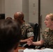 A group of U.S. Marines are seated around a conference table during what appears to be a briefing or meeting. A female Marine in MARPAT camouflage uniform is actively speaking and gesturing with her hand, appearing to be the primary presenter or discussant. Several other Marines in similar uniforms are seated nearby, with documents and a coffee cup visible on the table.