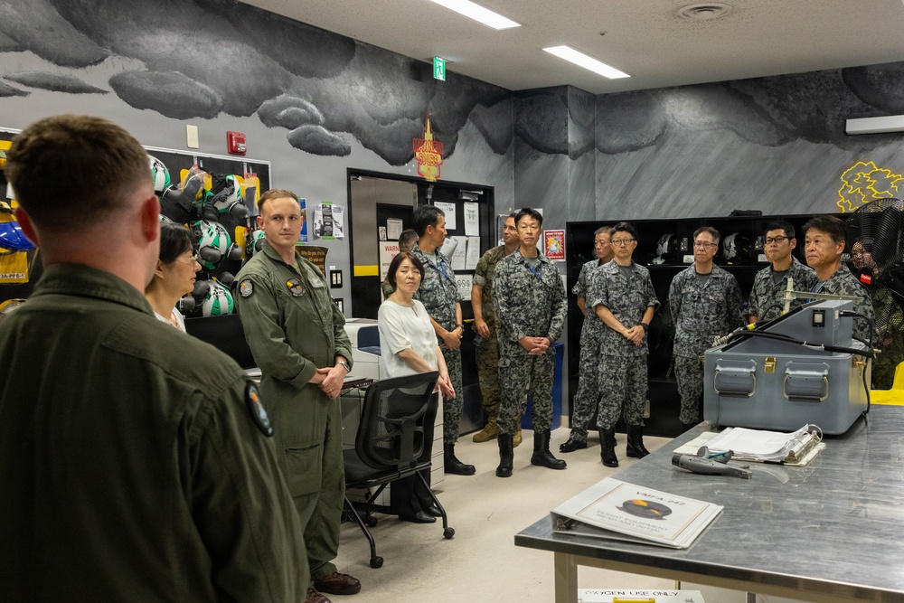 A briefing or orientation session is taking place in what appears to be a military aviation equipment room, likely at a U.S. Marine Corps installation. A mixed group of U.S. military personnel in olive flight suits and Japanese Self-Defense Force personnel in digital camouflage uniforms are gathered, suggesting a bilateral military exchange or tour. The room contains aviation gear including helmets and oxygen equipment, consistent with a flight equipment or life support section.
