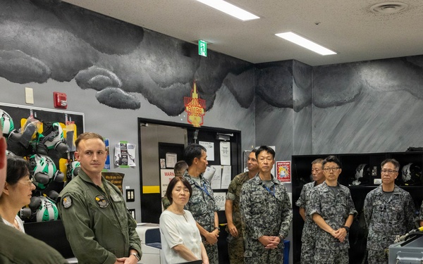 A briefing or orientation session is taking place in what appears to be a military aviation equipment room, likely at a U.S. Marine Corps installation. A mixed group of U.S. military personnel in olive flight suits and Japanese Self-Defense Force personnel in digital camouflage uniforms are gathered, suggesting a bilateral military exchange or tour. The room contains aviation gear including helmets and oxygen equipment, consistent with a flight equipment or life support section.