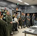 A briefing or orientation session is taking place in what appears to be a military aviation equipment room, likely at a U.S. Marine Corps installation. A mixed group of U.S. military personnel in olive flight suits and Japanese Self-Defense Force personnel in digital camouflage uniforms are gathered, suggesting a bilateral military exchange or tour. The room contains aviation gear including helmets and oxygen equipment, consistent with a flight equipment or life support section.