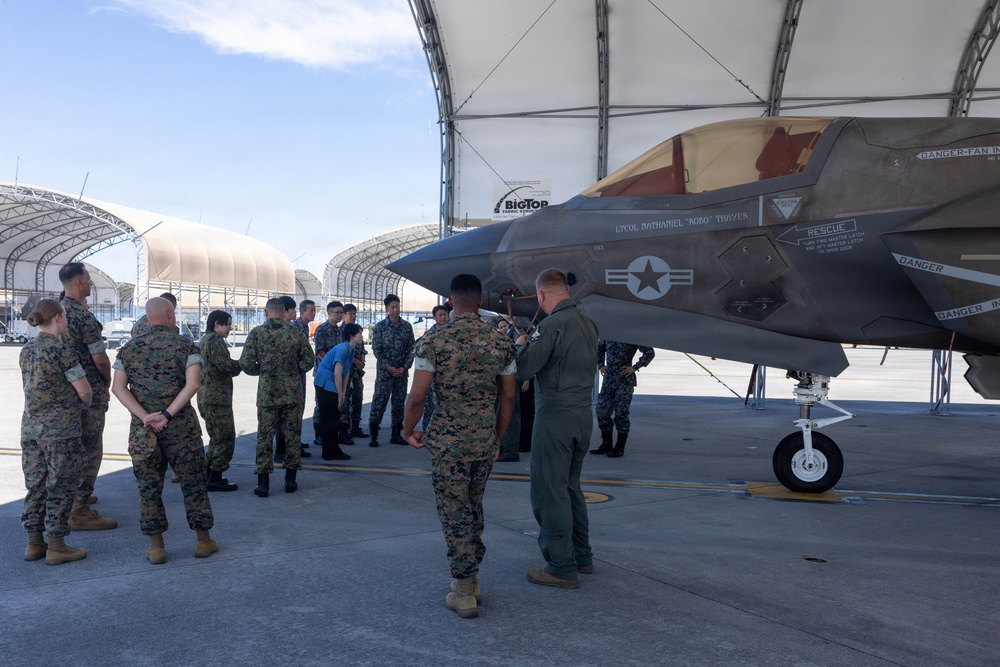 Military personnel from what appears to be two different armed forces are gathered on an airfield tarmac for a briefing or tour around an F-35 fighter jet. A pilot in a flight suit appears to be explaining or demonstrating features of the aircraft to the assembled group. The setting includes fabric hangar structures in the background, suggesting a forward operating base or temporary facility.