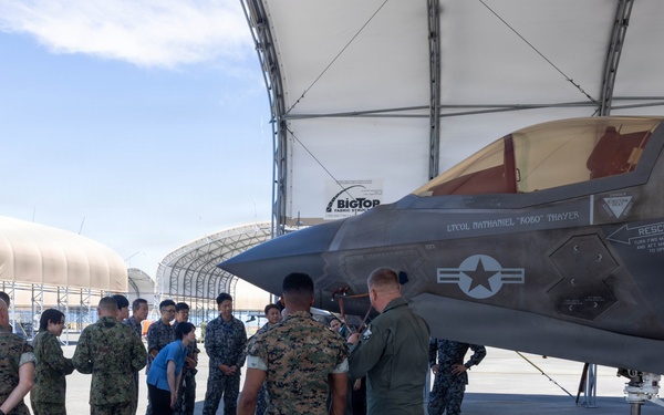 Military personnel from what appears to be two different armed forces are gathered on an airfield tarmac for a briefing or tour around an F-35 fighter jet. A pilot in a flight suit appears to be explaining or demonstrating features of the aircraft to the assembled group. The setting includes fabric hangar structures in the background, suggesting a forward operating base or temporary facility.