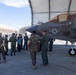 Military personnel from what appears to be two different armed forces are gathered on an airfield tarmac for a briefing or tour around an F-35 fighter jet. A pilot in a flight suit appears to be explaining or demonstrating features of the aircraft to the assembled group. The setting includes fabric hangar structures in the background, suggesting a forward operating base or temporary facility.