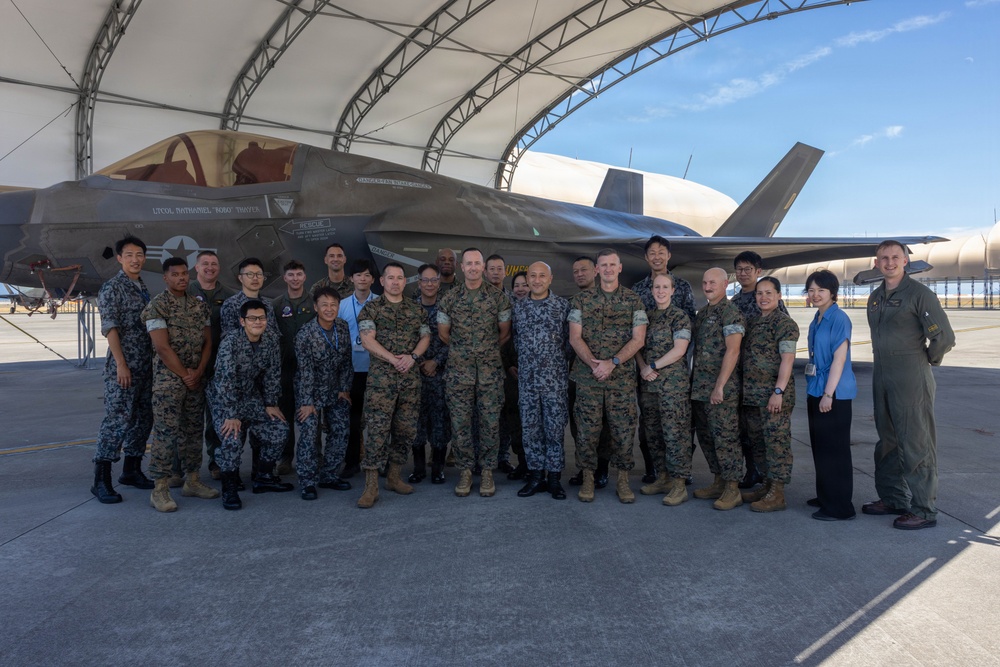 A group of approximately 25 military personnel pose for a formal group photograph in front of an F-35 fighter jet under a hangar shelter. The group includes members wearing multiple uniform types, suggesting a multinational or joint-service gathering. The setting appears to be a military airfield or flight line.