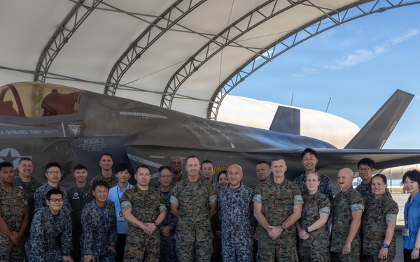 A group of approximately 25 military personnel pose for a formal group photograph in front of an F-35 fighter jet under a hangar shelter. The group includes members wearing multiple uniform types, suggesting a multinational or joint-service gathering. The setting appears to be a military airfield or flight line.