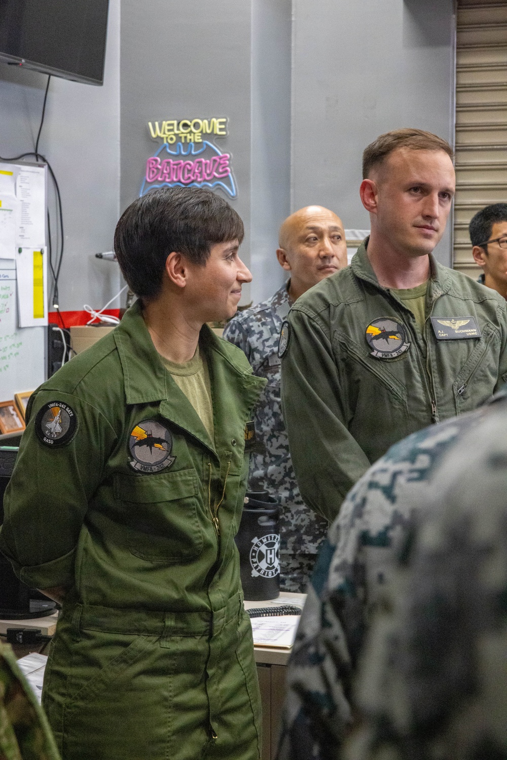 Military personnel in olive green flight suits are gathered in what appears to be a squadron ready room or operations space. Two foreground figures wear patches indicating VMFA-242 (a U.S. Marine Corps F/A-18 squadron), with the male figure's name tag reading 'A.J. Buchmann, Capt, USMC'. A neon sign reading 'Welcome to the Batcave' is visible in the background, a common informal nickname for squadron spaces.
