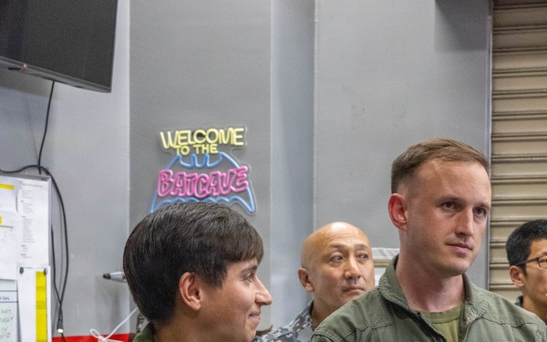 Military personnel in olive green flight suits are gathered in what appears to be a squadron ready room or operations space. Two foreground figures wear patches indicating VMFA-242 (a U.S. Marine Corps F/A-18 squadron), with the male figure's name tag reading 'A.J. Buchmann, Capt, USMC'. A neon sign reading 'Welcome to the Batcave' is visible in the background, a common informal nickname for squadron spaces.