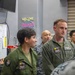 Military personnel in olive green flight suits are gathered in what appears to be a squadron ready room or operations space. Two foreground figures wear patches indicating VMFA-242 (a U.S. Marine Corps F/A-18 squadron), with the male figure's name tag reading 'A.J. Buchmann, Capt, USMC'. A neon sign reading 'Welcome to the Batcave' is visible in the background, a common informal nickname for squadron spaces.