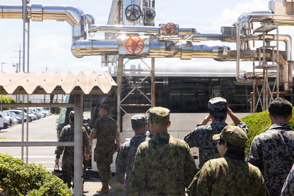 A group of military personnel in mixed uniforms are gathered near an industrial facility with large silver pipes and valves overhead. The soldiers appear to be on a tour or inspection visit, with some saluting and a bus visible in the background. The presence of multiple camouflage uniform styles suggests a joint military exercise or bilateral visit.