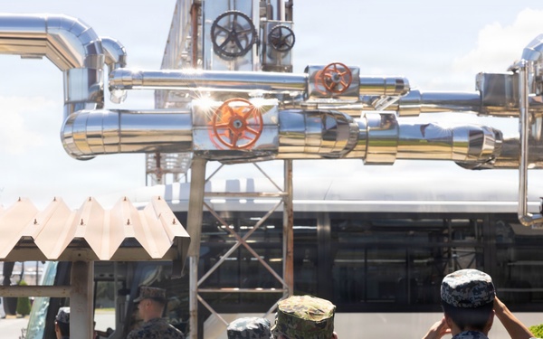 A group of military personnel in mixed uniforms are gathered near an industrial facility with large silver pipes and valves overhead. The soldiers appear to be on a tour or inspection visit, with some saluting and a bus visible in the background. The presence of multiple camouflage uniform styles suggests a joint military exercise or bilateral visit.
