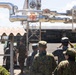 A group of military personnel in mixed uniforms are gathered near an industrial facility with large silver pipes and valves overhead. The soldiers appear to be on a tour or inspection visit, with some saluting and a bus visible in the background. The presence of multiple camouflage uniform styles suggests a joint military exercise or bilateral visit.
