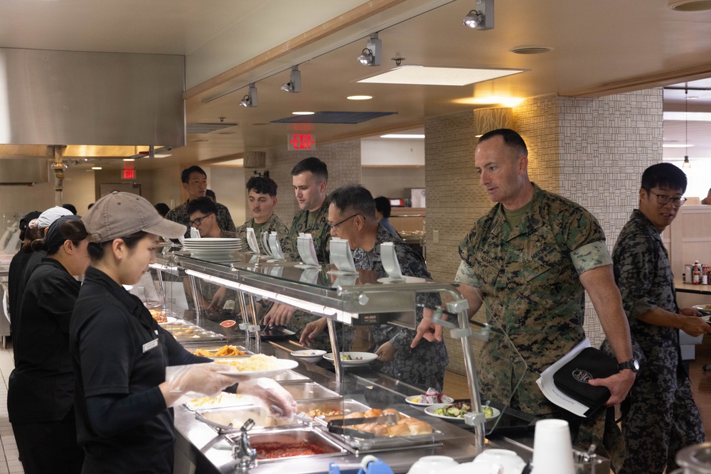 The image shows military personnel, appearing to be U.S. Marines in woodland MARPAT camouflage uniforms, going through a cafeteria-style chow hall or mess hall food service line. Civilian food service workers in black uniforms and caps are serving food from a steam table buffet line. The setting appears to be a military dining facility (DFAC), possibly on a joint or overseas installation given the mixed personnel visible.