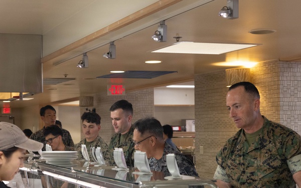 The image shows military personnel, appearing to be U.S. Marines in woodland MARPAT camouflage uniforms, going through a cafeteria-style chow hall or mess hall food service line. Civilian food service workers in black uniforms and caps are serving food from a steam table buffet line. The setting appears to be a military dining facility (DFAC), possibly on a joint or overseas installation given the mixed personnel visible.