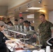 The image shows military personnel, appearing to be U.S. Marines in woodland MARPAT camouflage uniforms, going through a cafeteria-style chow hall or mess hall food service line. Civilian food service workers in black uniforms and caps are serving food from a steam table buffet line. The setting appears to be a military dining facility (DFAC), possibly on a joint or overseas installation given the mixed personnel visible.