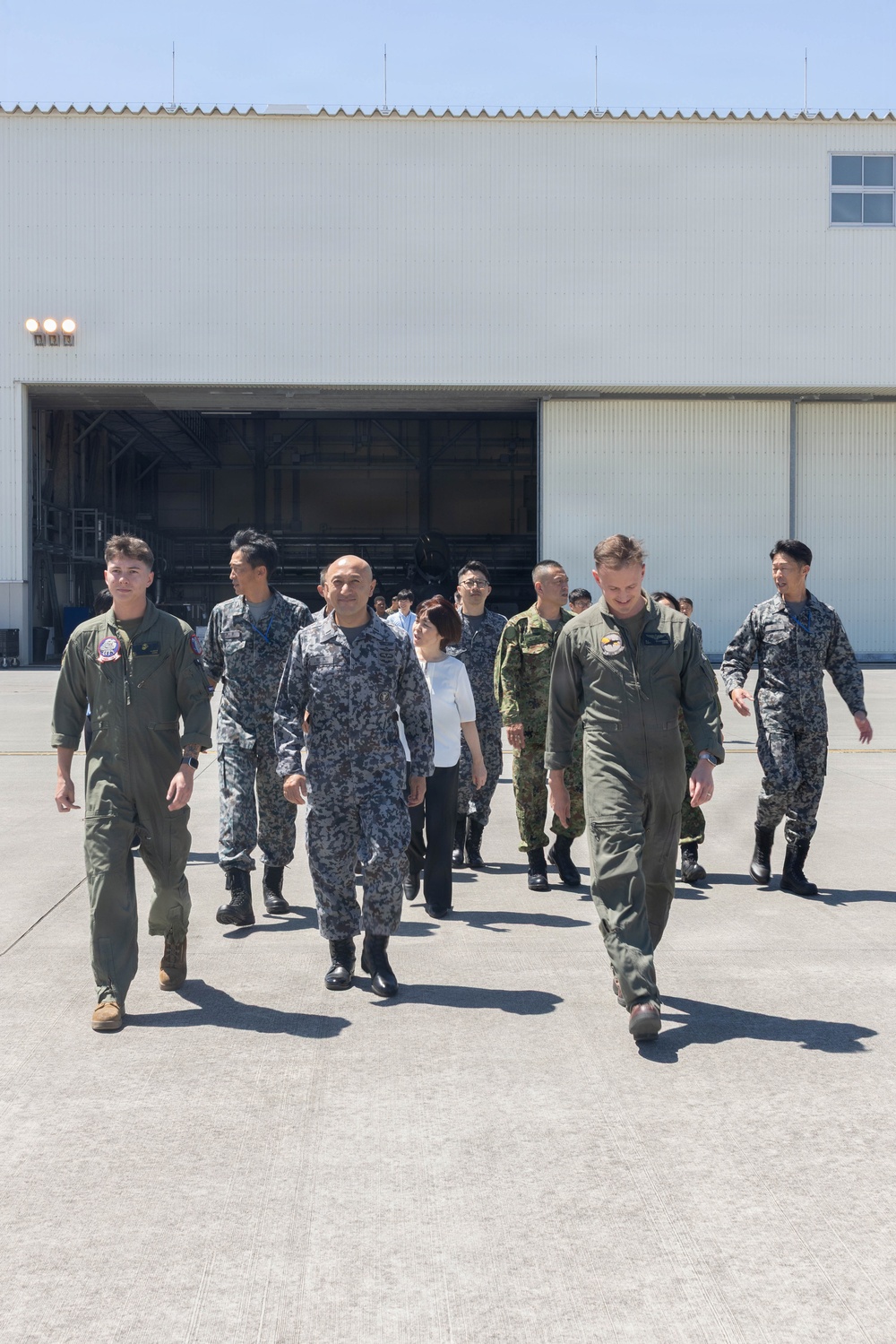 A group of military personnel and civilians walk across an airfield tarmac in front of a large aircraft hangar. The group includes individuals wearing olive green flight suits, blue-grey digital camouflage uniforms consistent with Japan Air Self-Defense Force (JASDF), green camouflage uniforms, and civilian attire. The setting appears to be a joint military base visit or tour conducted under clear, sunny conditions.
