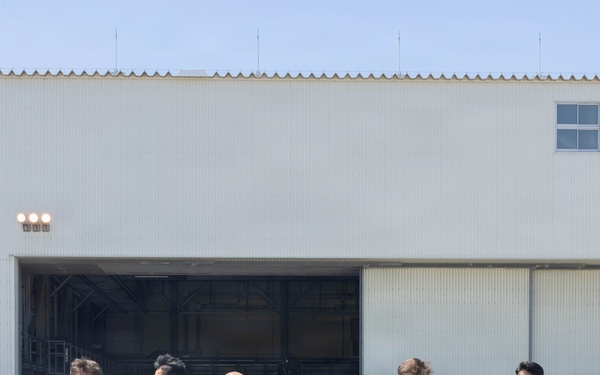 A group of military personnel and civilians walk across an airfield tarmac in front of a large aircraft hangar. The group includes individuals wearing olive green flight suits, blue-grey digital camouflage uniforms consistent with Japan Air Self-Defense Force (JASDF), green camouflage uniforms, and civilian attire. The setting appears to be a joint military base visit or tour conducted under clear, sunny conditions.