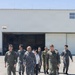 A group of military personnel and civilians walk across an airfield tarmac in front of a large aircraft hangar. The group includes individuals wearing olive green flight suits, blue-grey digital camouflage uniforms consistent with Japan Air Self-Defense Force (JASDF), green camouflage uniforms, and civilian attire. The setting appears to be a joint military base visit or tour conducted under clear, sunny conditions.