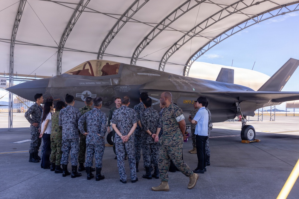 A group of military personnel from multiple nations are gathered under an open aircraft shelter examining an F-35 fighter jet marked with 'VMFA-2' on its tail, indicating a U.S. Marine Corps Fighter Attack Squadron. Personnel wearing at least two distinct camouflage patterns — U.S. Marine MARPAT and a blue/grey digital pattern consistent with Japanese Self-Defense Force uniforms — are present alongside civilians. The setting appears to be a military airfield, likely during a bilateral or multilateral exchange or demonstration event.