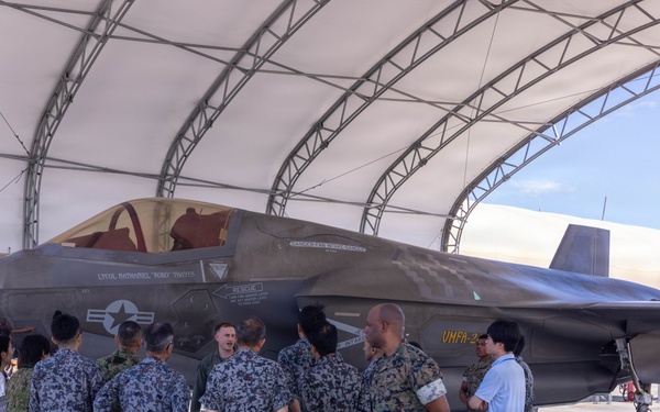A group of military personnel from multiple nations are gathered under an open aircraft shelter examining an F-35 fighter jet marked with 'VMFA-2' on its tail, indicating a U.S. Marine Corps Fighter Attack Squadron. Personnel wearing at least two distinct camouflage patterns — U.S. Marine MARPAT and a blue/grey digital pattern consistent with Japanese Self-Defense Force uniforms — are present alongside civilians. The setting appears to be a military airfield, likely during a bilateral or multilateral exchange or demonstration event.
