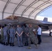 A group of military personnel from multiple nations are gathered under an open aircraft shelter examining an F-35 fighter jet marked with 'VMFA-2' on its tail, indicating a U.S. Marine Corps Fighter Attack Squadron. Personnel wearing at least two distinct camouflage patterns — U.S. Marine MARPAT and a blue/grey digital pattern consistent with Japanese Self-Defense Force uniforms — are present alongside civilians. The setting appears to be a military airfield, likely during a bilateral or multilateral exchange or demonstration event.