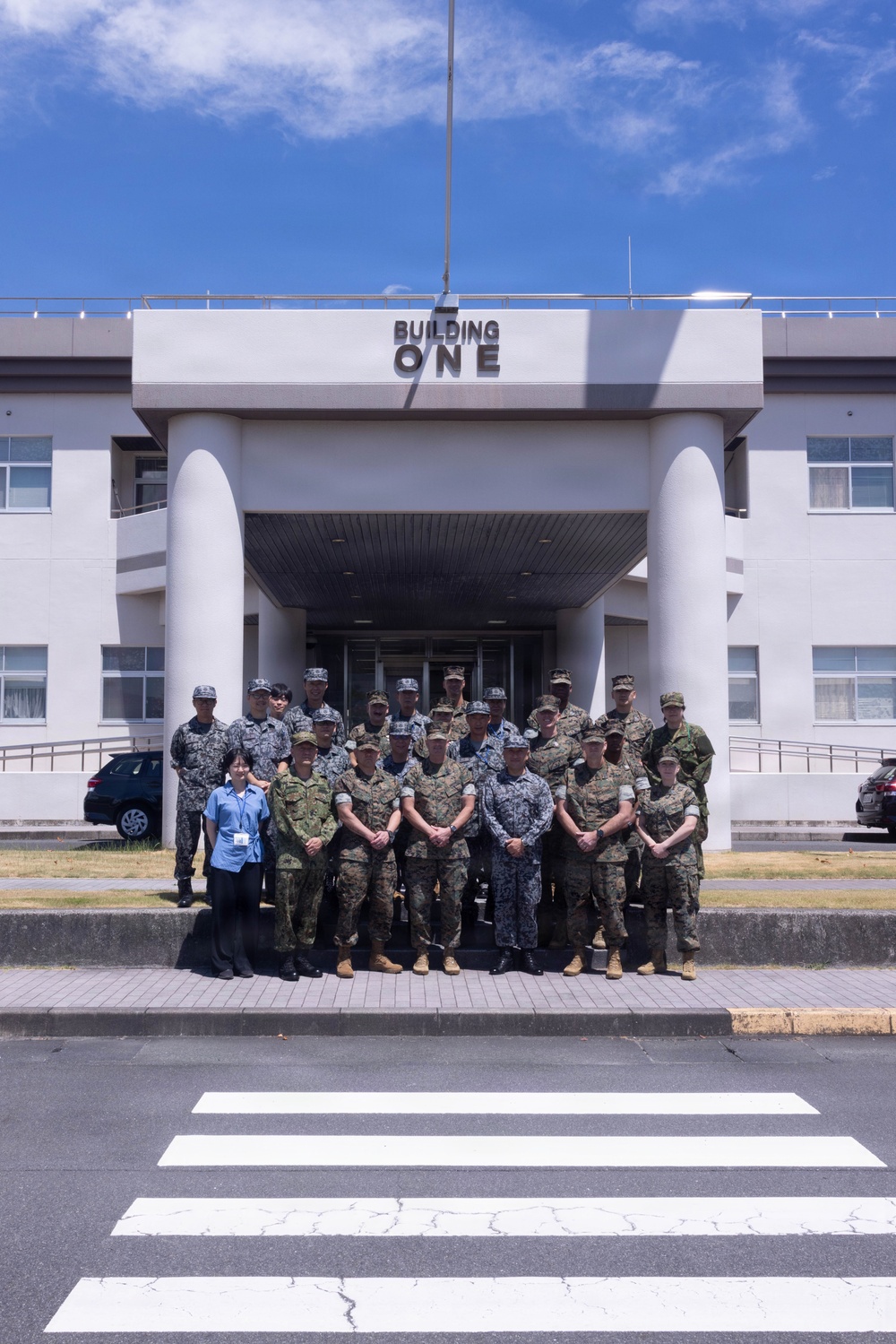 A group of approximately 20-25 military personnel and one civilian pose for a formal group photograph in front of a building labeled 'BUILDING ONE.' The personnel are wearing multiple distinct camouflage uniform patterns, suggesting a multinational or joint-service gathering. The photo is taken from across a pedestrian crosswalk on a clear, sunny day.