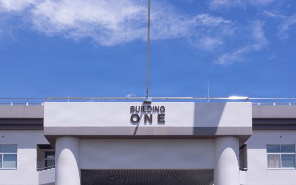 A group of approximately 20-25 military personnel and one civilian pose for a formal group photograph in front of a building labeled 'BUILDING ONE.' The personnel are wearing multiple distinct camouflage uniform patterns, suggesting a multinational or joint-service gathering. The photo is taken from across a pedestrian crosswalk on a clear, sunny day.
