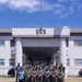 A group of approximately 20-25 military personnel and one civilian pose for a formal group photograph in front of a building labeled 'BUILDING ONE.' The personnel are wearing multiple distinct camouflage uniform patterns, suggesting a multinational or joint-service gathering. The photo is taken from across a pedestrian crosswalk on a clear, sunny day.