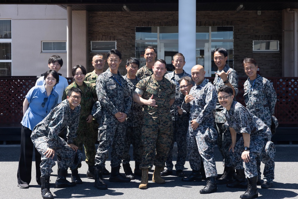 A group photo of approximately 15 individuals, predominantly wearing Japanese Air Self-Defense Force (JASDF) digital camouflage uniforms, posing with what appears to be a U.S. Marine in woodland MARPAT camouflage. The group is gathered in front of a building that appears to be a military facility, suggesting a joint military exchange or cooperative event. Two civilians in casual attire are also present in the background.
