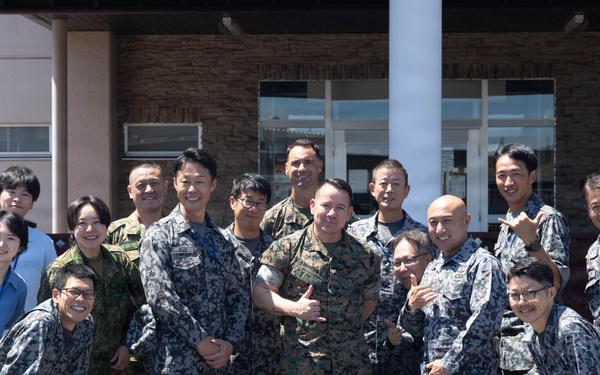A group photo of approximately 15 individuals, predominantly wearing Japanese Air Self-Defense Force (JASDF) digital camouflage uniforms, posing with what appears to be a U.S. Marine in woodland MARPAT camouflage. The group is gathered in front of a building that appears to be a military facility, suggesting a joint military exchange or cooperative event. Two civilians in casual attire are also present in the background.