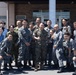A group photo of approximately 15 individuals, predominantly wearing Japanese Air Self-Defense Force (JASDF) digital camouflage uniforms, posing with what appears to be a U.S. Marine in woodland MARPAT camouflage. The group is gathered in front of a building that appears to be a military facility, suggesting a joint military exchange or cooperative event. Two civilians in casual attire are also present in the background.