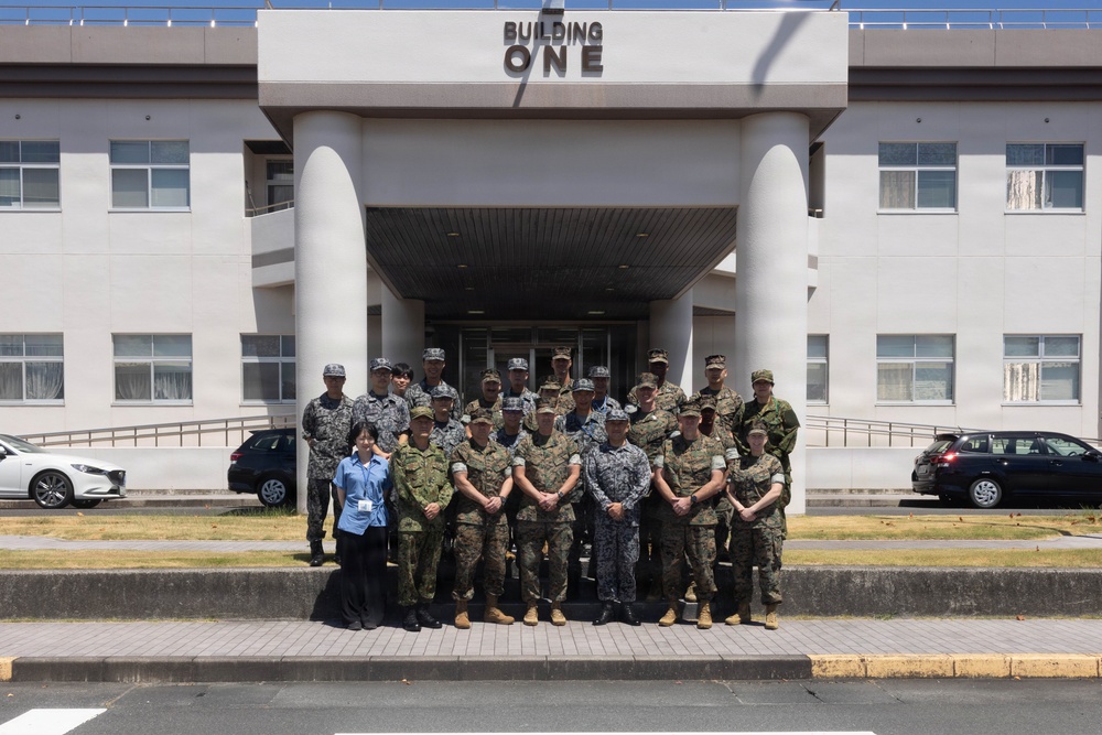 A group of approximately 20 military personnel in various camouflage uniforms pose for a formal group photograph in front of a building labeled 'Building One.' The group includes individuals wearing different uniform patterns, suggesting a multinational or multi-branch military gathering. One civilian in a blue shirt is also present among the group.
