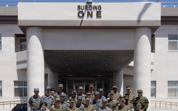 A group of approximately 20 military personnel in various camouflage uniforms pose for a formal group photograph in front of a building labeled 'Building One.' The group includes individuals wearing different uniform patterns, suggesting a multinational or multi-branch military gathering. One civilian in a blue shirt is also present among the group.
