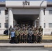 A group of approximately 20 military personnel in various camouflage uniforms pose for a formal group photograph in front of a building labeled 'Building One.' The group includes individuals wearing different uniform patterns, suggesting a multinational or multi-branch military gathering. One civilian in a blue shirt is also present among the group.