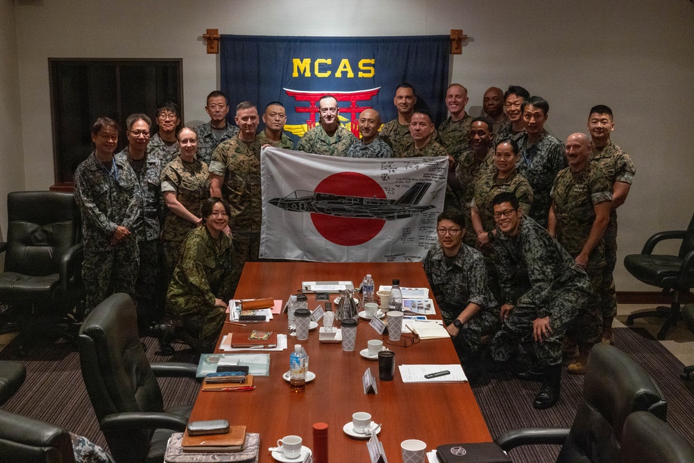 A group of military personnel from what appears to be the United States Marine Corps and Japan Self-Defense Force are gathered in a conference room at MCAS (Marine Corps Air Station) for a group photo. Two individuals in the center are holding a Japanese flag (Hinomaru) with a military aircraft silhouette and handwritten signatures on it. The setting suggests a bilateral military exchange or liaison meeting.