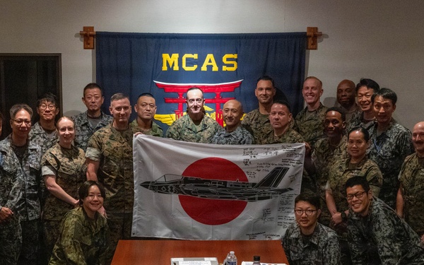 A group of military personnel from what appears to be the United States Marine Corps and Japan Self-Defense Force are gathered in a conference room at MCAS (Marine Corps Air Station) for a group photo. Two individuals in the center are holding a Japanese flag (Hinomaru) with a military aircraft silhouette and handwritten signatures on it. The setting suggests a bilateral military exchange or liaison meeting.