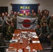 A group of military personnel from what appears to be the United States Marine Corps and Japan Self-Defense Force are gathered in a conference room at MCAS (Marine Corps Air Station) for a group photo. Two individuals in the center are holding a Japanese flag (Hinomaru) with a military aircraft silhouette and handwritten signatures on it. The setting suggests a bilateral military exchange or liaison meeting.