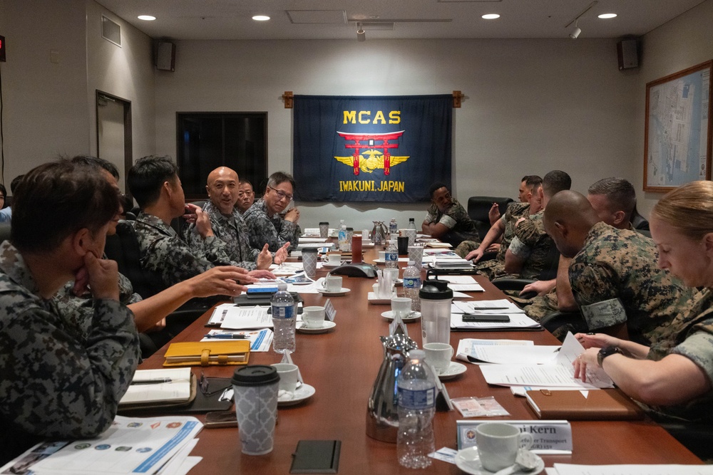 A joint military meeting is taking place at MCAS Iwakuni, Japan, with personnel from both the U.S. Marine Corps and Japanese Self-Defense Forces seated around a conference table. The MCAS Iwakuni flag is prominently displayed on the wall behind the participants. The meeting appears to involve bilateral coordination or planning, with documents, water bottles, and coffee cups spread across the table.