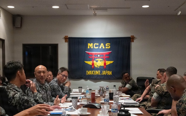 A joint military meeting is taking place at MCAS Iwakuni, Japan, with personnel from both the U.S. Marine Corps and Japanese Self-Defense Forces seated around a conference table. The MCAS Iwakuni flag is prominently displayed on the wall behind the participants. The meeting appears to involve bilateral coordination or planning, with documents, water bottles, and coffee cups spread across the table.
