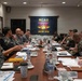 A joint military meeting is taking place at MCAS Iwakuni, Japan, with personnel from both the U.S. Marine Corps and Japanese Self-Defense Forces seated around a conference table. The MCAS Iwakuni flag is prominently displayed on the wall behind the participants. The meeting appears to involve bilateral coordination or planning, with documents, water bottles, and coffee cups spread across the table.