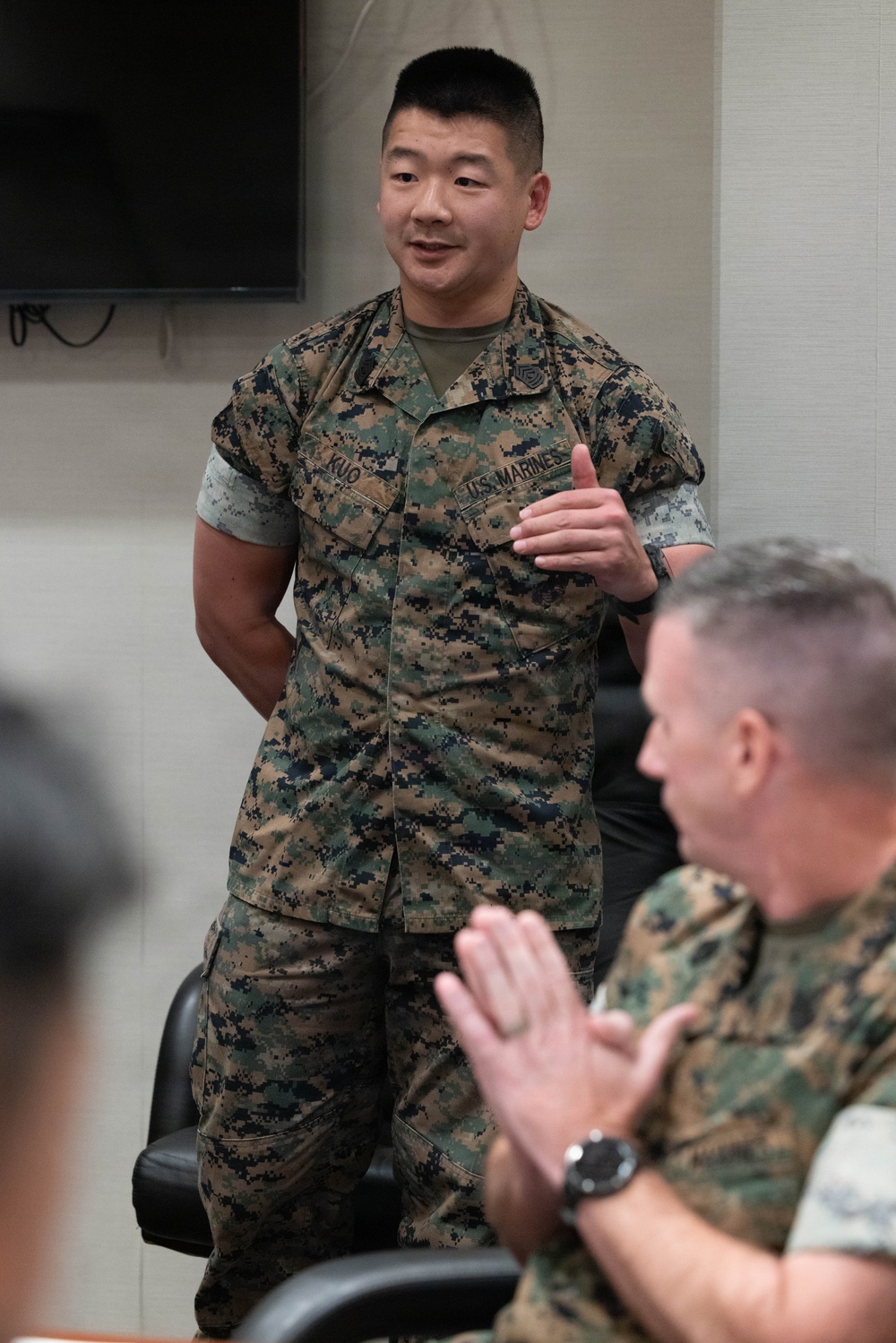 A U.S. Marine in MARPAT digital camouflage uniform with the name tape 'KUO' and 'U.S. MARINES' tape is standing and speaking or presenting in what appears to be an indoor briefing room. A second Marine in the foreground is seated and appears to be applauding. The setting suggests a formal or semi-formal military meeting or recognition event.