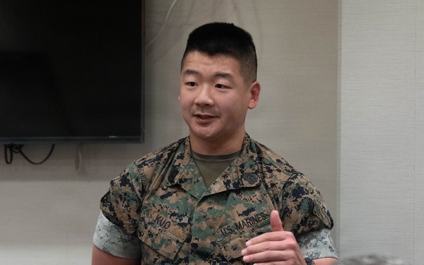 A U.S. Marine in MARPAT digital camouflage uniform with the name tape 'KUO' and 'U.S. MARINES' tape is standing and speaking or presenting in what appears to be an indoor briefing room. A second Marine in the foreground is seated and appears to be applauding. The setting suggests a formal or semi-formal military meeting or recognition event.
