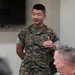 A U.S. Marine in MARPAT digital camouflage uniform with the name tape 'KUO' and 'U.S. MARINES' tape is standing and speaking or presenting in what appears to be an indoor briefing room. A second Marine in the foreground is seated and appears to be applauding. The setting suggests a formal or semi-formal military meeting or recognition event.