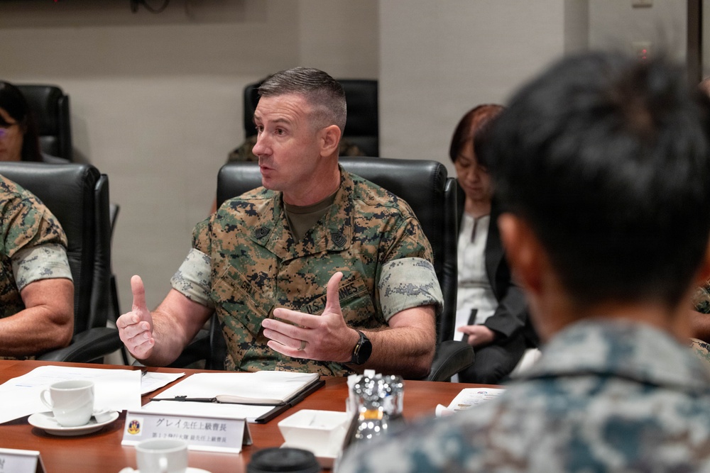 A U.S. Marine in MARPAT camouflage uniform is speaking at a formal meeting table, gesturing with both hands. A nameplate in Japanese characters is visible on the table in front of him, indicating an official bilateral or multinational military meeting. Other attendees, including a civilian woman and personnel in different uniforms, are seated around the table.