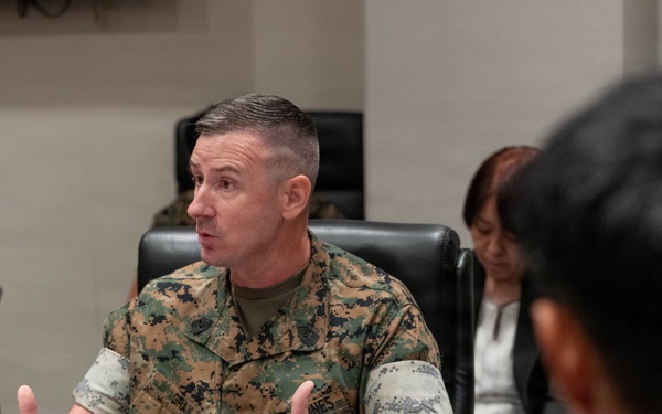 A U.S. Marine in MARPAT camouflage uniform is speaking at a formal meeting table, gesturing with both hands. A nameplate in Japanese characters is visible on the table in front of him, indicating an official bilateral or multinational military meeting. Other attendees, including a civilian woman and personnel in different uniforms, are seated around the table.