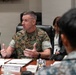 A U.S. Marine in MARPAT camouflage uniform is speaking at a formal meeting table, gesturing with both hands. A nameplate in Japanese characters is visible on the table in front of him, indicating an official bilateral or multinational military meeting. Other attendees, including a civilian woman and personnel in different uniforms, are seated around the table.