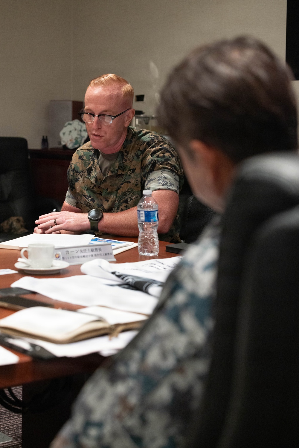 A U.S. Marine in MARPAT camouflage uniform is seated at a conference table, appearing to speak during a formal meeting. A name placard in Japanese script is visible on the table in front of him, along with documents, a water bottle, and a tea cup. A second individual in the foreground, partially visible and out of focus, appears to be wearing a camouflage uniform as well.