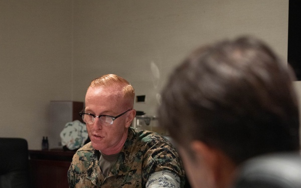 A U.S. Marine in MARPAT camouflage uniform is seated at a conference table, appearing to speak during a formal meeting. A name placard in Japanese script is visible on the table in front of him, along with documents, a water bottle, and a tea cup. A second individual in the foreground, partially visible and out of focus, appears to be wearing a camouflage uniform as well.